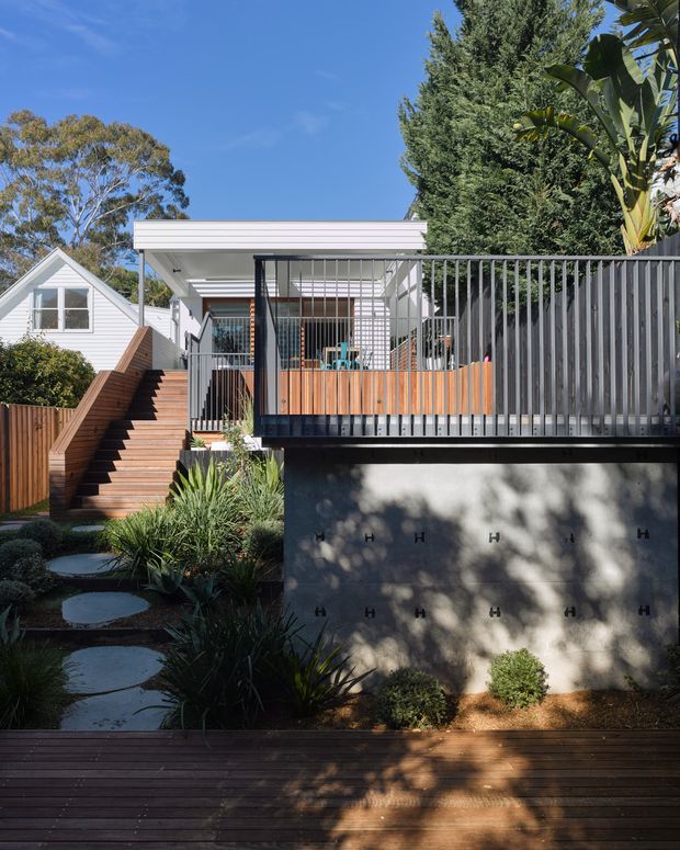 A bright exterior view of a contemporary elevated dwelling and landscaped garden. A timber stair rises to a covered upper-level deck with timber balustrading, slim metal screens and a white flat-roof canopy. A dark concrete retaining wall supports the deck, while stepping stones, drought-tolerant planting and a timber deck occupy the foreground. A neighbouring light weatherboard gabled structure is visible at left.
