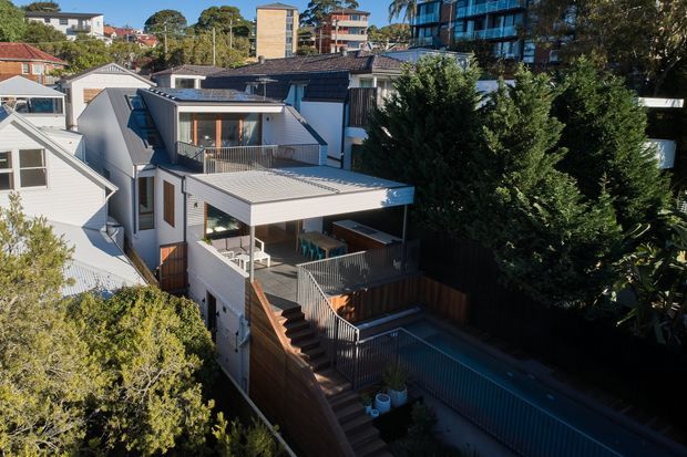 Elevated exterior view of a contemporary multi-level residential house in a dense suburban setting. The home features light cladding, dark pitched roofing, a low-pitched metal-roofed extension, upper-level glazing, multiple timber decks and stairs, and a narrow rectangular swimming pool enclosed by fencing and mature vegetation. Apartment buildings and neighbouring homes are visible in the background.
