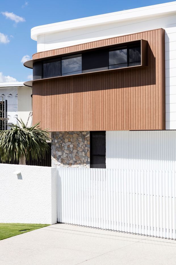 Exterior photograph of a contemporary multi-level residence with a rounded white roofline, warm vertical timber cladding, dark horizontal window band, and a natural irregular-stone feature wall. A white slatted fence and gate enclose the front boundary, with a small landscaped planting area and driveway in the foreground.