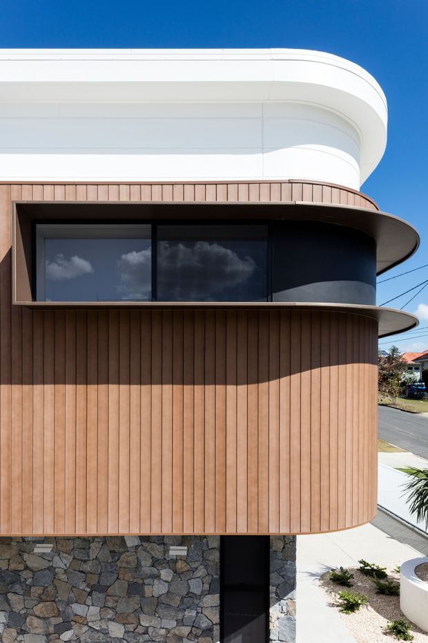Close-up exterior view of a contemporary multi-level building with strongly rounded, aerodynamic forms. The facade combines warm vertical timber-look cladding, dark glazed ribbon windows, a smooth white curved upper volume, and a rugged irregular stone base. A narrow dark entry is visible below, with a suburban street and neighbouring homes in the background.