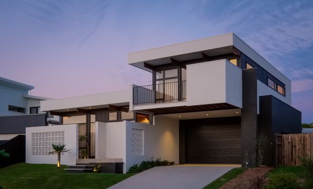 Exterior dusk view of a contemporary multi-storey residence with a strong geometric composition, flat rooflines, white rendered volumes, dark cladding, extensive glazing, a first-floor balcony, integrated garage, decorative breeze-block screens, and a landscaped front approach.