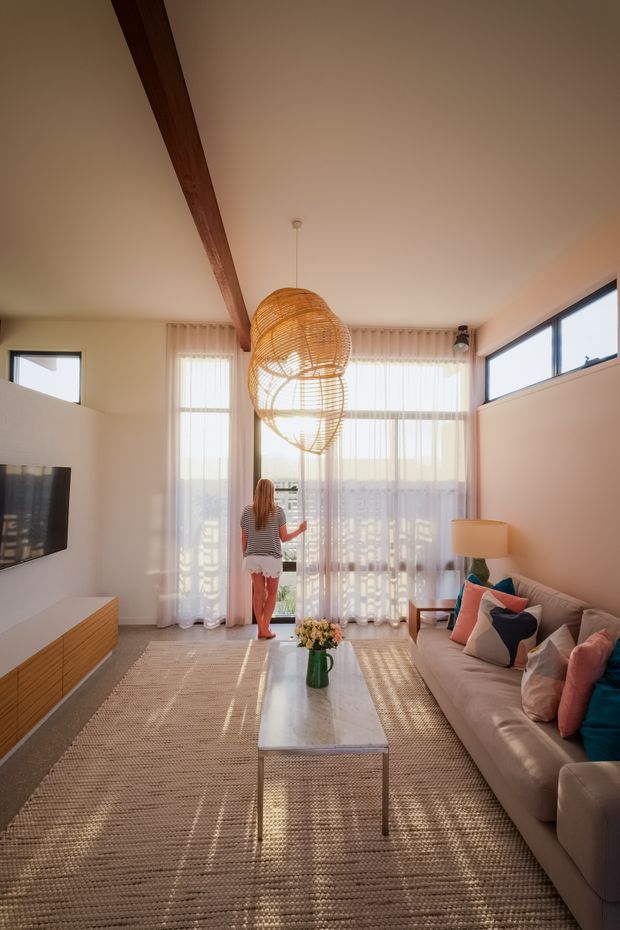 Bright contemporary living room with a warm neutral palette, large floor-to-ceiling windows dressed in sheer curtains, a timber ceiling beam, woven pendant light, beige sectional sofa with colourful cushions, textured natural-fibre rug, wood media console and a slim coffee table. The room opens toward a balcony or exterior view through the glazing.
