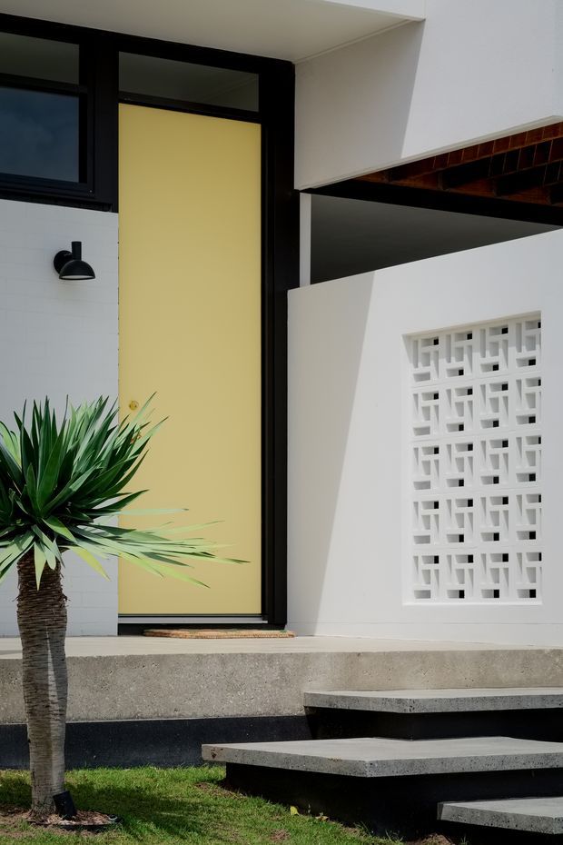 A close exterior view of a modern residential entrance. The facade combines white rendered walls, black-framed glazing and a pale yellow front door. A geometric white breeze-block screen provides ventilation beside the entry, while floating concrete steps lead up from a small landscaped garden with a palm-like plant. Dark timber is visible beneath the overhead roof or canopy.