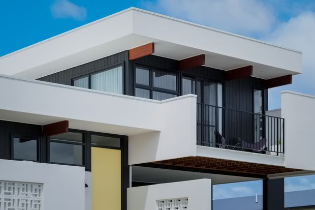 Exterior view of a contemporary multi-level residence with crisp white rendered volumes, dark vertical cladding, extensive black-framed glazing, projecting flat roof planes, and a small upper balcony with metal railing. Timber-toned structural accents and brickwork are visible beneath the balcony. Decorative breeze-block screens appear at the lower level.