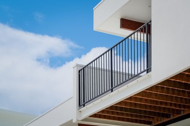 Close-up exterior architectural view of a contemporary multi-level building, focused on a projecting balcony with slim black metal balustrades, white rendered walls, and a warm timber-lined soffit beneath the overhang. The image is taken from below against a blue sky, highlighting clean geometric forms and cantilevered construction.