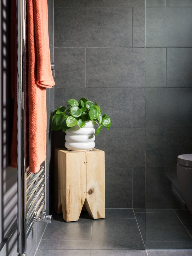 A modern bathroom corner finished in large dark grey tiles, with a chrome heated towel rail beside a window and coral towel. A leafy green plant in a white rounded planter sits on a natural timber stump pedestal, while the edge of a toilet is visible on the right. The space has a refined, high-contrast, contemporary feel with natural wood and greenery softening the dark tiled surfaces.