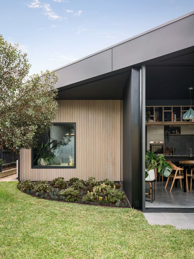 Exterior view of a contemporary single-storey home with a low-pitched roof, dark metal framing, vertical timber wall cladding, and expansive glazed openings. A landscaped garden borders the facade, while the open glazing reveals a dining and kitchen area inside.