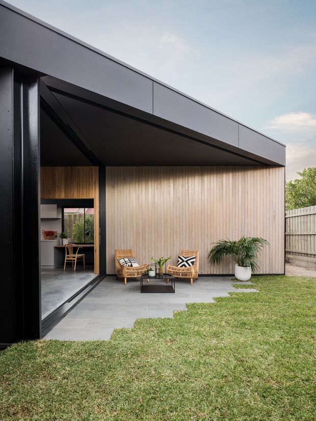 Contemporary single-storey pavilion-style home extension with a low-pitched flat roof, dark metal framing and cladding, warm vertical timber wall panelling, and large open glazing. A paved outdoor seating area with woven armchairs and potted plants fronts a lawn, while an interior dining or living space is visible through the opening.