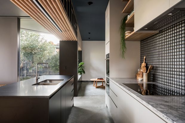 Contemporary galley kitchen with a large island, integrated sink and tap, dark cabinetry, white stone benchtops, and a glossy black tiled splashback. A timber slatted ceiling feature runs above the island, while full-height glazing brings in natural light and views of greenery. The kitchen includes open shelving, concealed appliances, and a compact seating nook beyond.