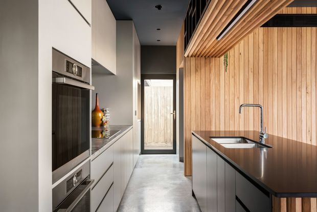 A narrow contemporary galley kitchen with tall white integrated appliances and cabinetry on the left, and a dark stone island with sink and tap on the right. Warm vertical timber wall panelling and timber ceiling battens contrast with the polished concrete floor and dark ceiling. A glazed door at the rear provides natural light and access to an outdoor area.