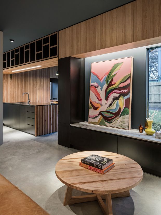 A sophisticated open-plan interior featuring a dark, minimalist kitchen with black cabinetry, vertical timber wall panelling, and a slatted timber island end. A large abstract artwork rests on a black built-in display cabinet beneath warm timber overhead joinery. The space has a polished concrete floor, dark ceiling, integrated linear lighting, a round timber coffee table, and a large window overlooking greenery.