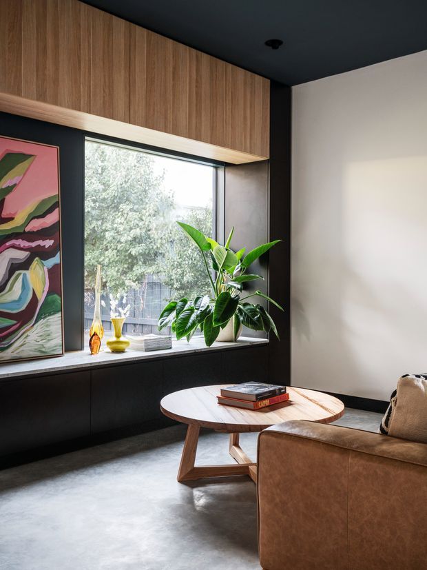 A sophisticated living room with a large window, dark built-in cabinetry, warm timber overhead joinery, a polished concrete floor, and a brown leather sofa. A sculptural timber coffee table, indoor plant, artwork, and decorative objects add warmth and colour. The room has a moody, refined contemporary character with strong natural light.
