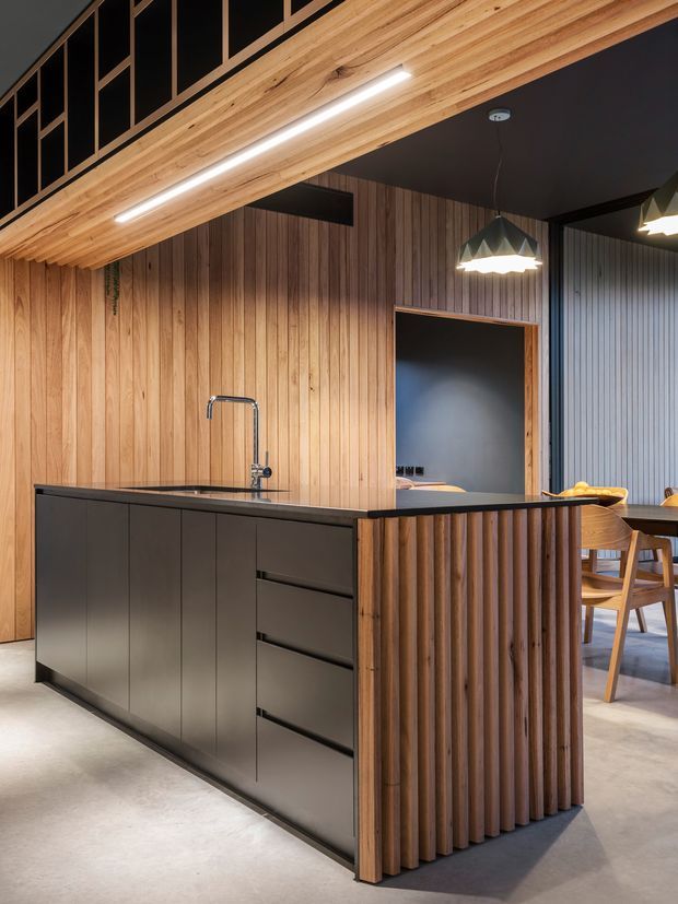 A contemporary open-plan kitchen featuring a large dark matte island with integrated sink and tap, vertical timber wall panelling, and a fluted timber end panel. The ceiling combines warm timber lining with black sections and integrated linear lighting. A dining area with timber chairs is visible beyond the island, with a dark pendant light above it.
