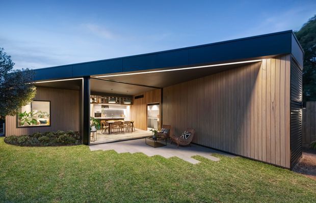 Exterior twilight view of a contemporary single-storey pavilion-style home with a dark flat roof, extensive open glazing, warm vertical timber wall cladding, and integrated linear lighting. The covered opening reveals a dining area and kitchen, while a landscaped lawn, stepping stones, planting, and outdoor seating occupy the foreground.
