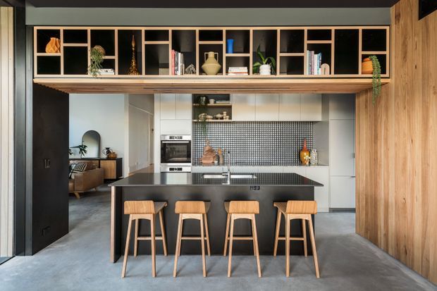 A contemporary open-plan kitchen viewed from the living area, featuring a large black island with four timber bar stools, white cabinetry, a small square-tile backsplash, integrated oven, and extensive open timber shelving with books and decorative objects. Black-framed openings, dark cabinetry, timber wall panelling, and a polished concrete floor create a refined industrial-modern interior. A living room is visible through the opening on the left.