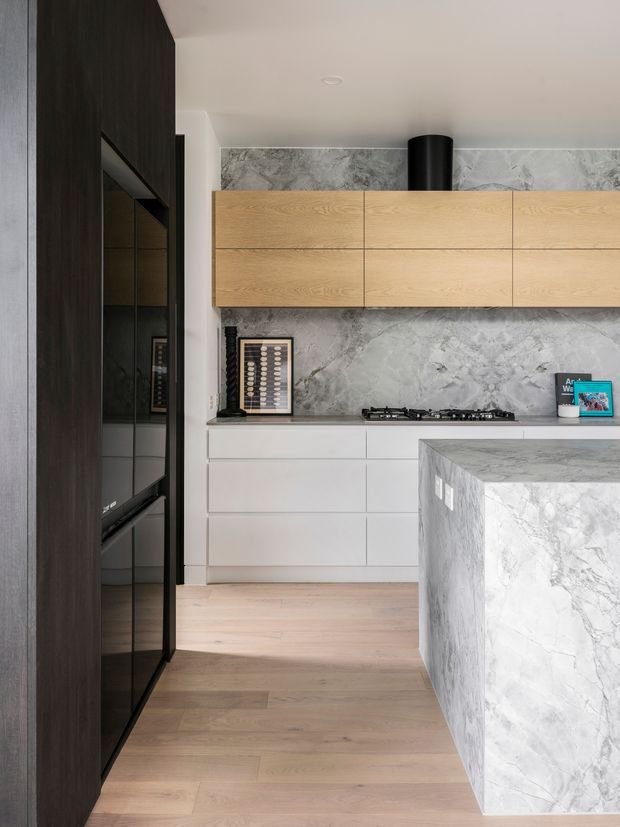 Contemporary kitchen with light timber overhead cabinetry, white lower drawers, a dramatic grey marble-look splashback and matching waterfall island, integrated black appliance cabinetry, pale timber flooring, and a black cylindrical rangehood. The compact space has a refined, high-contrast palette with warm wood accents.