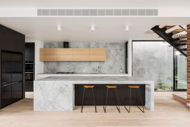 A contemporary open-plan kitchen viewed straight on, featuring a large marble-look island with integrated seating, dark lower cabinetry, warm timber wall cabinetry, a full-height veined stone splashback, and black appliances. A glazed opening and exposed brick stair structure are visible at the right, with pale timber flooring throughout.