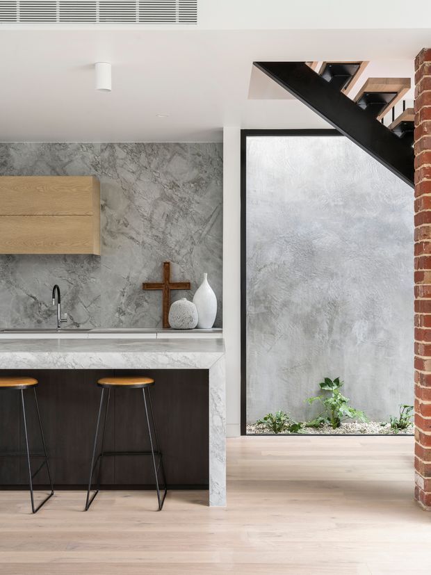 A contemporary kitchen viewed beside an internal courtyard or lightwell. The kitchen features a dark timber island with a pale marble-look benchtop, grey stone-look splashback, timber wall cabinetry, black tapware, and two slim stools. Pale timber flooring extends toward a textured grey courtyard wall with planting. A black steel stair structure and exposed red brick wall add strong architectural contrast.