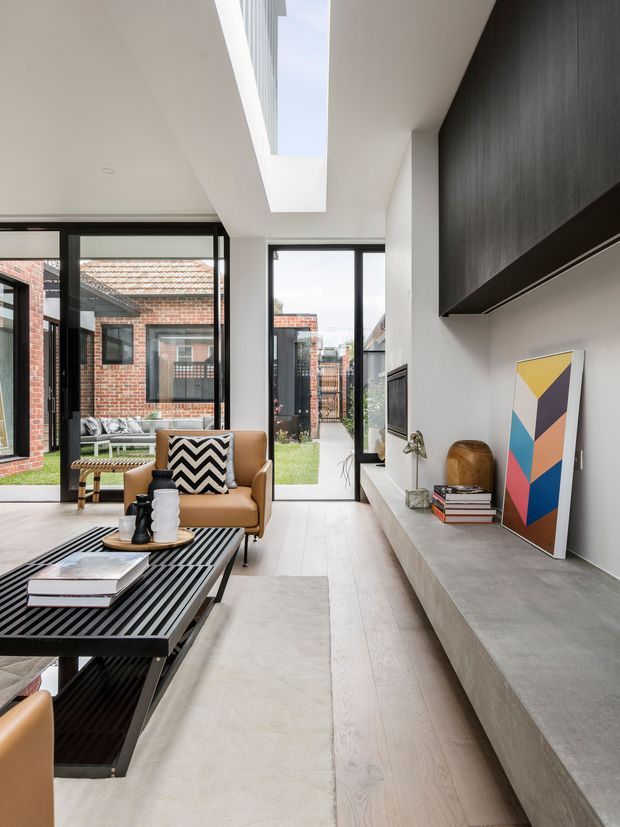 A bright, contemporary open-plan living room with a tan leather armchair, black slatted coffee table, and long built-in concrete-look media bench and cabinetry. Full-height black-framed glazing opens toward a small courtyard, while a rectangular skylight brings additional daylight into the white interior. The room combines pale timber flooring, white walls, dark joinery, and colourful geometric artwork.