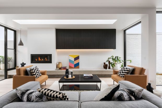 Bright, contemporary living room with a large light-grey sectional sofa in the foreground, two tan leather armchairs, a black coffee table, and a wall-mounted dark timber entertainment cabinet. The room features white walls, broad black-framed glazing, a linear fireplace, a skylight, indoor plants, and monochrome patterned cushions with a colourful artwork accent.