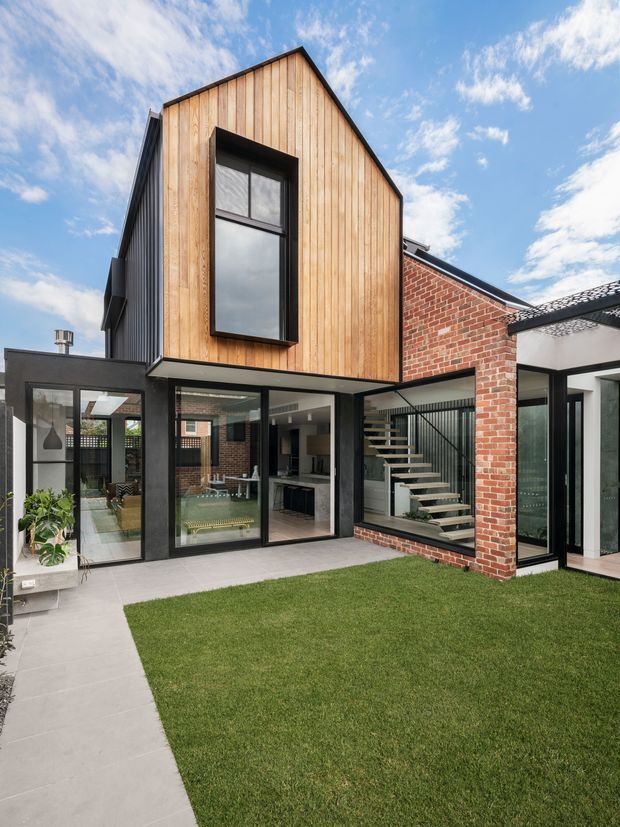 Contemporary multi-level residential extension viewed from a landscaped courtyard. The facade combines vertical timber weatherboarding, exposed red brick, black-framed glazing and a projecting dark metal roof edge. Large glazed openings reveal a bright interior with a floating staircase, while a paved path and lawn occupy the foreground.