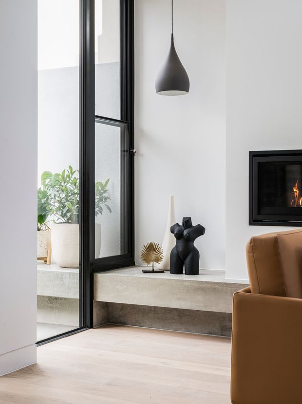 A refined contemporary living room with a tall black-framed glazed door or window, pale timber flooring, white walls, and a built-in concrete fireplace hearth. A black suspended pendant light, sculptural decor, indoor plants, and a caramel leather armchair create a warm minimalist composition.