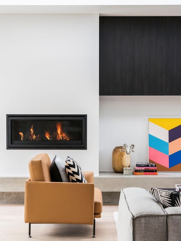 Contemporary living room with a built-in linear fireplace set into a smooth white wall, dark timber overhead cabinetry, and a long stone or concrete-look hearth ledge. A tan leather armchair and grey sectional sofa frame the room, with graphic cushions, sculptural decor, books, and colourful geometric artwork adding contrast.