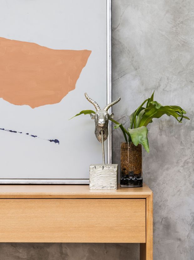 Close-up interior vignette featuring a light timber sideboard with clean, flush-front joinery against a textured grey plaster wall. Decorative objects and a potted plant sit on top, alongside a large abstract artwork with a peach and black painted composition. The styling is understated and contemporary with natural materials and muted tones.