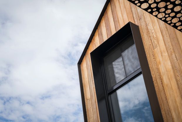 Close-up upward view of a contemporary building facade clad in vertical warm timber boards, with a large black-framed window projecting from the wall. The underside of the roof or eave features a distinctive circular-patterned soffit, with sky reflected in the glazing.