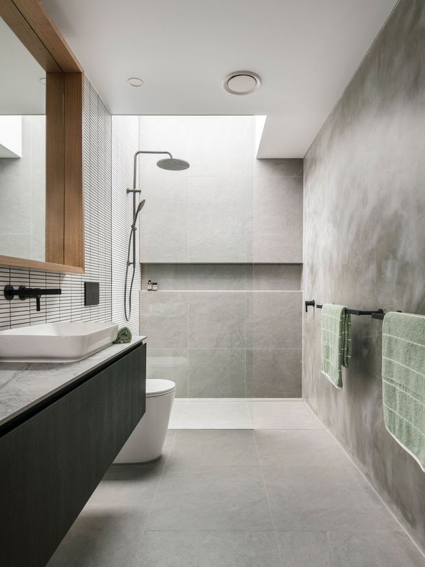 A contemporary bathroom with a walk-in shower, wall-mounted rain shower and handheld fixture, floating dark vanity with white vessel basin, wall-mounted toilet, large grey floor and wall tiles, a tiled accent wall, timber-framed mirror cabinetry, and a textured plaster-like wall with mint green towels.