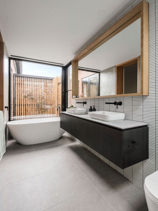 Contemporary bathroom with a freestanding white bathtub beside full-height glazing, a large timber-framed mirrored cabinet, twin vessel basins, matte black wall-mounted tapware, dark floating vanity cabinetry, white horizontal tile wall lining, and large grey floor tiles. A timber privacy fence is visible outside.