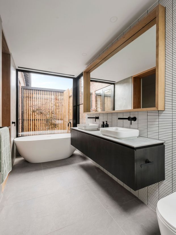 Contemporary bathroom with a freestanding white bathtub beside full-height glazing, a double floating vanity with white vessel basins, black wall-mounted tapware, a large timber-framed mirror, and fine white linear wall tiles. The room has large grey floor tiles, dark cabinetry, and a private timber-fenced courtyard visible through the window.