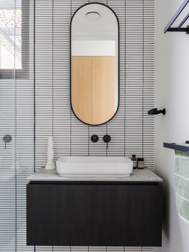 Contemporary bathroom vanity with a white vessel basin, dark timber cabinetry, pale stone benchtop, and a wall clad in narrow horizontal white tiles with dark grout. A large rounded-rectangle black-framed mirror hangs above the basin, with matte black wall-mounted tapware and accessories. A glass shower enclosure is visible to the left.