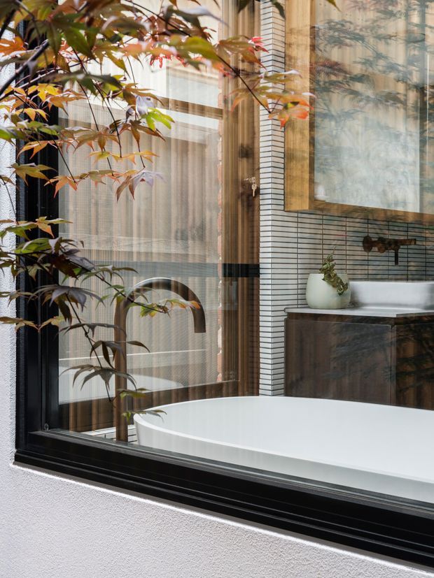 A refined bathroom viewed through a large black-framed window, featuring a freestanding white bathtub, dark timber vanity cabinetry, white vertical mosaic wall tiles, a framed wall artwork or mirror, and a small potted plant. Exterior foliage is reflected or visible across the glazing, creating a layered indoor-outdoor view.