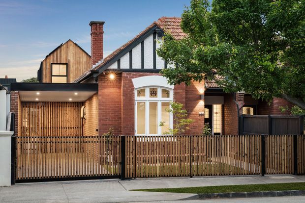 Street-facing view of a renovated brick residence with a prominent gabled front facade, white Tudor-style half-timbering, a tiled pitched roof, tall chimney, and a warm timber-slatted carport or entry structure. A vertical timber front fence and gate enclose the landscaped frontage.