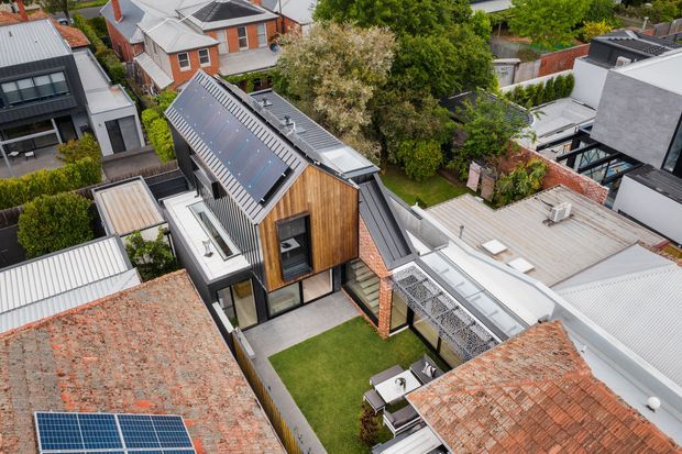 Aerial view of a compact urban residential property surrounded by neighbouring homes. The central contemporary house combines a steep pitched metal roof, vertical timber cladding, dark-framed glazing, exposed brick walls and a small landscaped courtyard. Rooftop elements, covered outdoor areas and solar panels are visible across the surrounding properties.