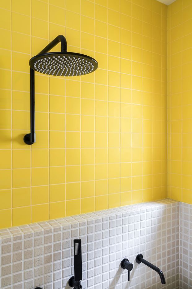 Close-up view of a vibrant bathroom shower and bath area. The walls are clad in glossy yellow square tiles, transitioning to a band of small pale grey mosaic tiles around the lower section. A matte black rainfall showerhead, hand shower, and bath tapware provide strong contrast against the bright tiled surfaces.