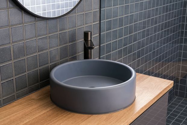 A close-up of a contemporary bathroom vanity featuring a round matte dark-grey vessel basin, black tapware, and a timber benchtop. The walls and floor are finished in small dark grey square tiles, with a partial round mirror visible above.