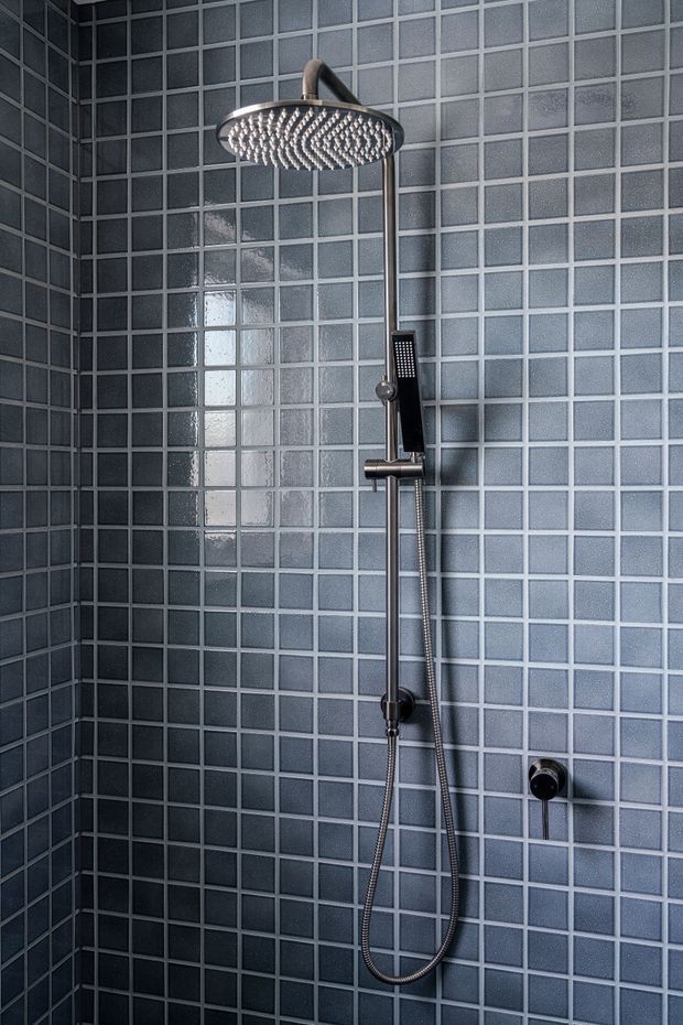 Close-up view of a modern walk-in shower enclosure with small square blue-grey wall tiles, dark grout, and brushed metal shower hardware including a large rain showerhead, handheld shower, rail, and controls.