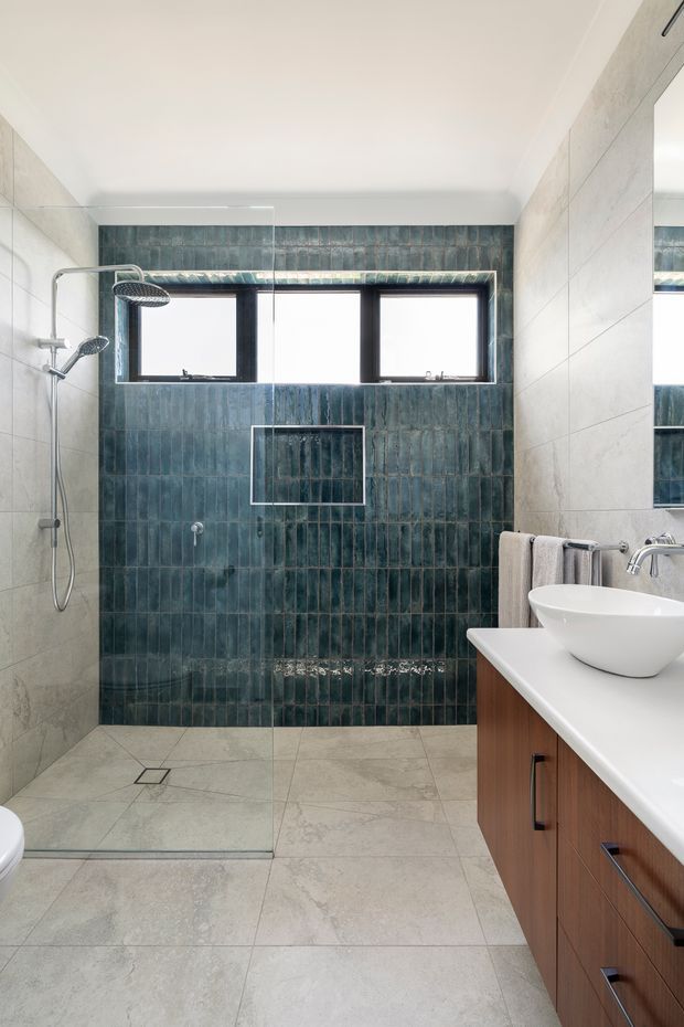 A bright contemporary bathroom with a walk-in glass shower, wall-mounted rainfall and handheld shower fixtures, dark blue-green vertical tile feature wall, pale large-format floor and side-wall tiles, and a timber-veneer vanity with a white vessel basin. A horizontal window provides natural light, and a mirrored cabinet or wall reflection is visible at right.