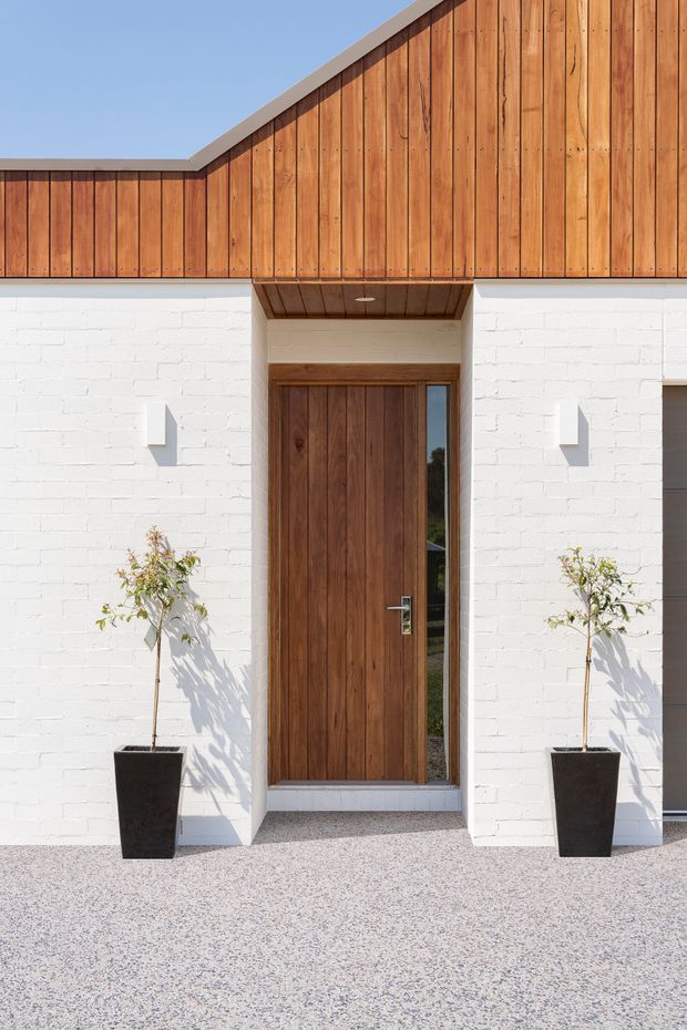 Front entrance of a contemporary single-storey building with a white-painted brick facade, vertically clad warm timber gable and matching timber entry door. The recessed doorway has a narrow glazed sidelight and timber-lined soffit, with two black planters flanking the entrance and a light aggregate-paved forecourt.