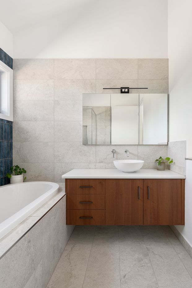 Bright contemporary bathroom with large light-grey stone-look wall and floor tiles, a white bathtub, floating mid-tone timber vanity with white benchtop, vessel basin and wall-mounted tapware. Mirrored wall cabinetry, a black linear light, and small potted greenery provide functional and decorative details. A dark blue tiled feature strip is visible along the left side.