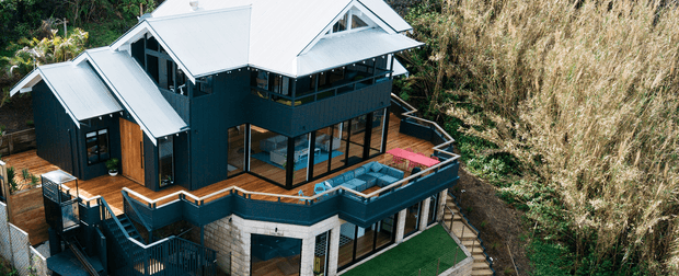 Aerial view of a multi-level contemporary house set among dense vegetation. The home has dark blue-green cladding, extensive glazing, multiple angular pitched roof forms in light grey metal, timber decks and balustrades, and several outdoor terraces including a rooftop-style entertaining area with seating.