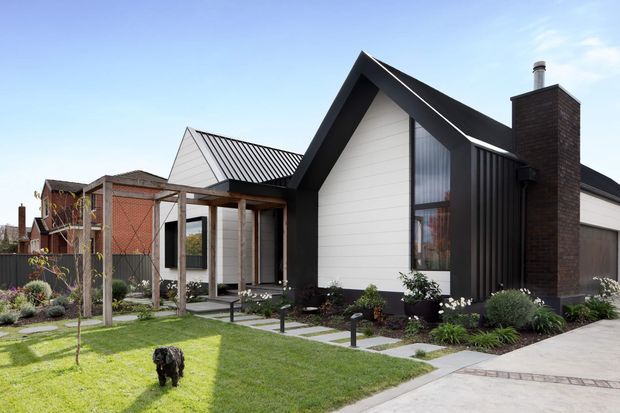 Exterior view of a contemporary single-storey residence with a steep gabled roof, black vertical cladding and trim, white horizontal wall cladding, and a prominent dark brick chimney. The home has a covered timber-framed entry or outdoor passage, landscaped front garden, paved stepping stones, and a concrete driveway. A neighbouring house is visible in the background.