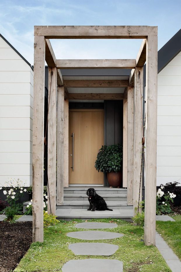 Front exterior view of a contemporary residential entry. A weathered timber portal frames a timber front door and covered approach, with broad tiled steps, irregular concrete stepping stones, lawn and planted garden beds. The facade uses white horizontal cladding with dark pitched roof edges. A small dog sits on the entry steps.