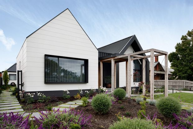 Exterior view of a contemporary residential home with a steep gabled white-clad volume, dark standing-seam metal roofs, black-framed glazing, and a weathered timber pergola over the entry terrace. The landscaped front garden includes clipped shrubs, ornamental planting, stepping-stone paths, and a fenced suburban setting.