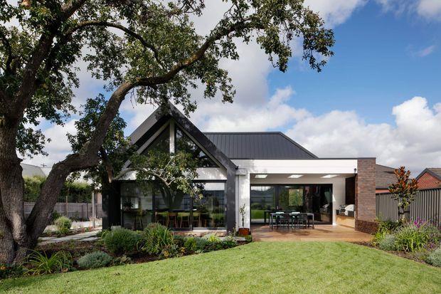 Exterior view of a contemporary single-storey home set in a landscaped suburban garden. The house features a prominent dark gabled glazed pavilion, a low dark metal roof extending over a covered outdoor dining area, white rendered walls, and a warm brick side wall. Large windows and sliding glass openings connect the living areas to the garden and patio.