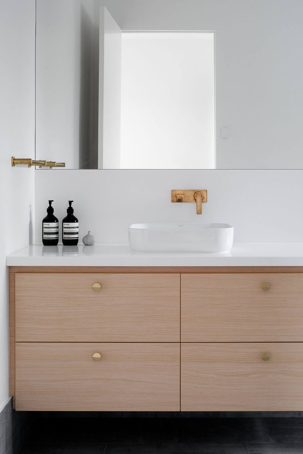 A minimalist bathroom vanity photographed straight on. The room features a floating light timber drawer vanity with small brass pulls, a white solid-surface benchtop, a white vessel basin, and a wall-mounted brass tap. A large mirrored cabinet or mirror spans the upper wall, with white and pale grey surfaces and black flooring creating a restrained, contemporary scheme.