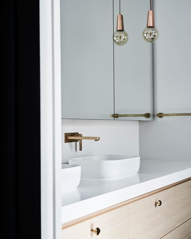 Close-up view of a refined bathroom vanity with a white vessel basin, wall-mounted brass tapware, pale stone or solid-surface benchtop, and light timber-grain drawers. Mirrored cabinetry with brass hardware occupies the upper wall, while two copper-toned pendant lights hang in front. The palette is light, clean, and softly warm.