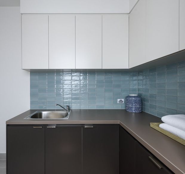 Compact contemporary kitchen or kitchenette with a U-shaped dark cabinetry layout, stainless-steel sink, matte dark benchtops, glossy pale blue subway-tile splashback, and streamlined white overhead cabinets. The space is clean and minimalist, with a small decorative container and folded towels on the counter.